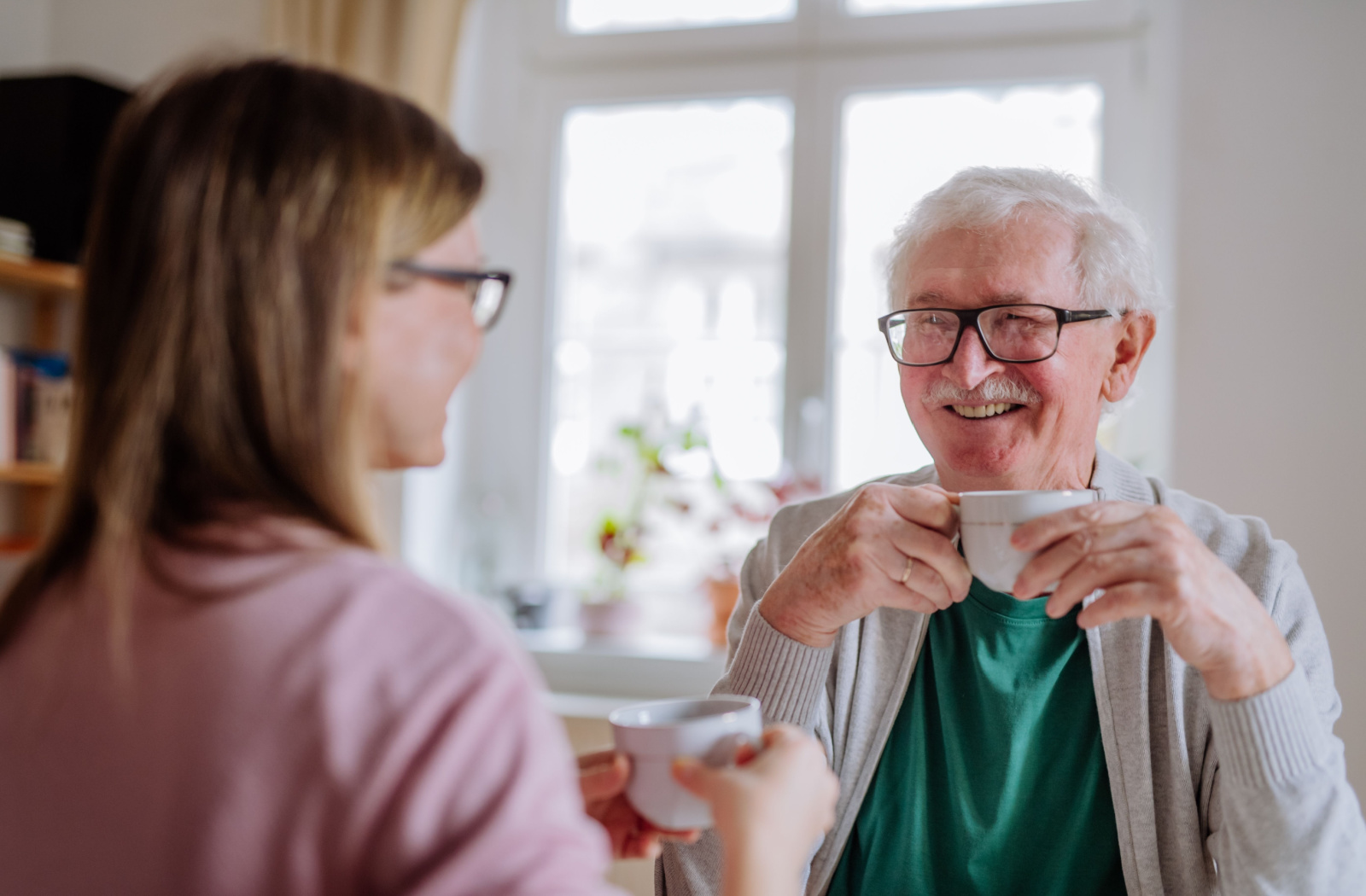 A senior father and his daughter sitting on a table, smiling and talking to each other while holding a cup of tea.
