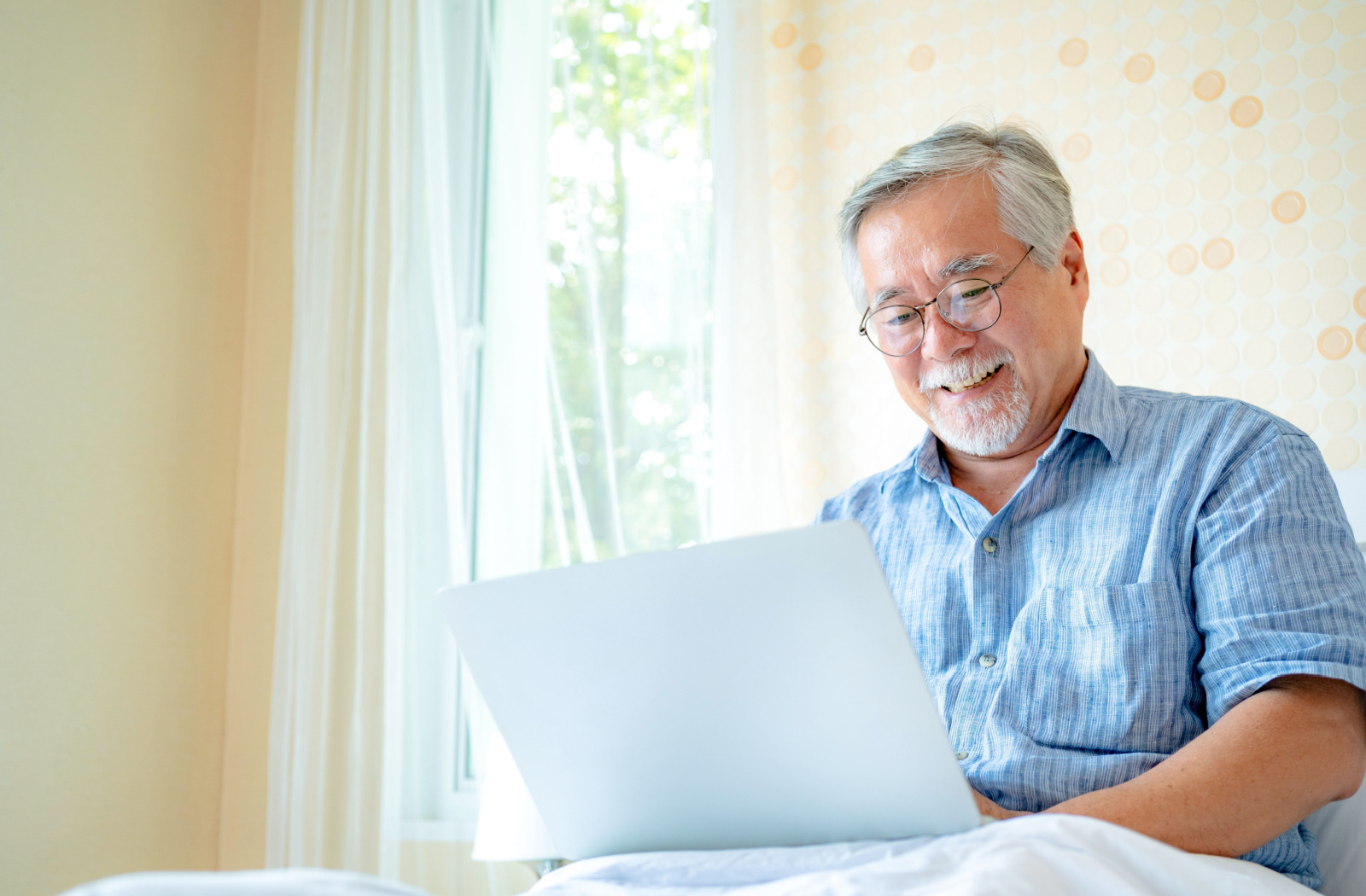 A senior man wearing a button-up shirt and glasses, sitting down and smiling while using a laptop.