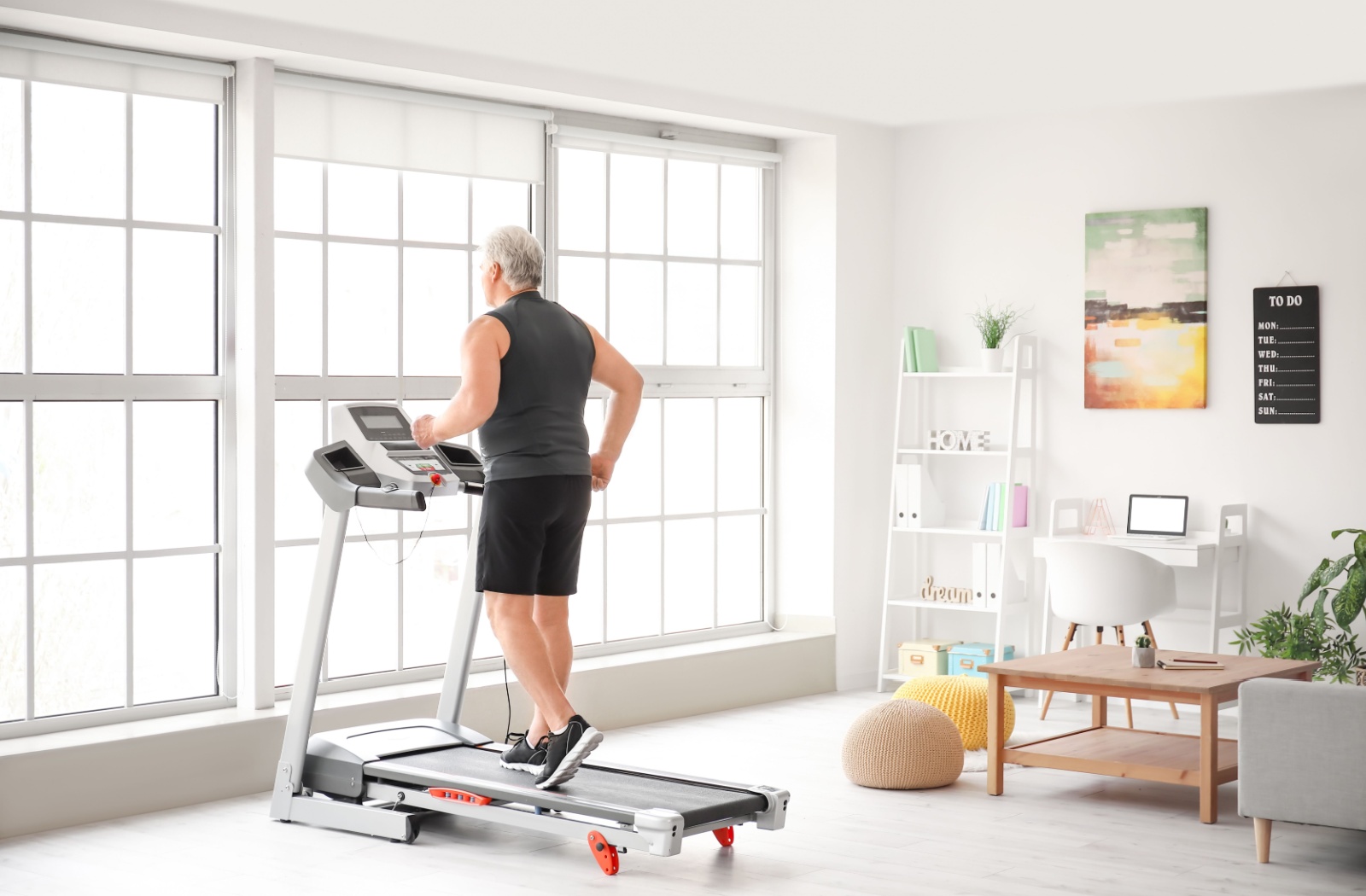 An older man walking on a folding treadmill in a bright and neatly organized room.