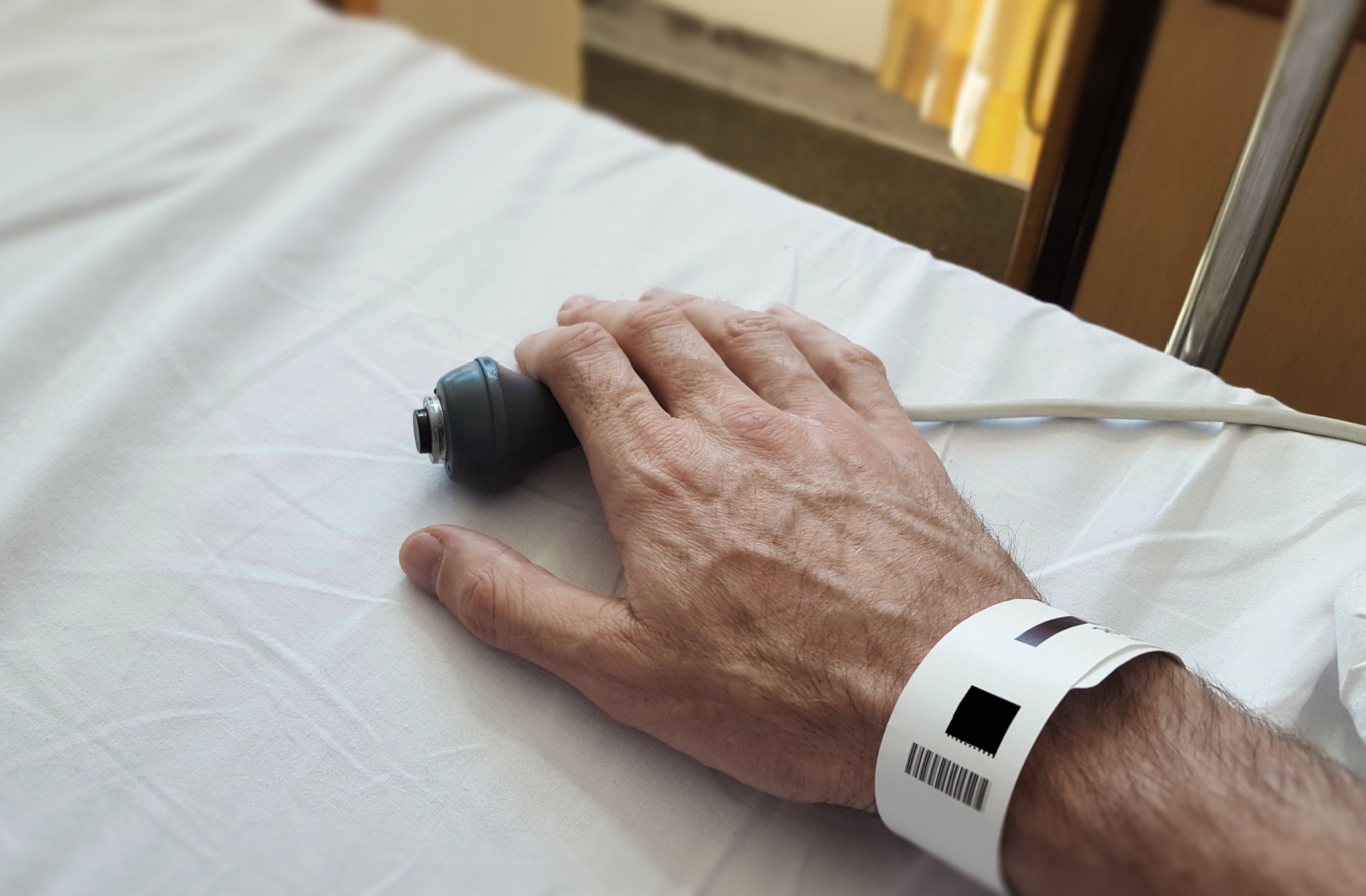 An older adult in a hospital bed holds the call button for assistance.
