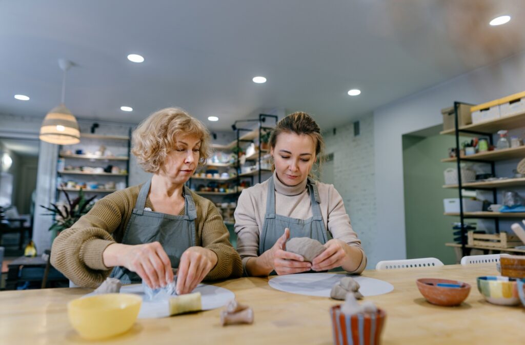An older adult carefully follows the instructions of a pottery teacher to learn a new skill.