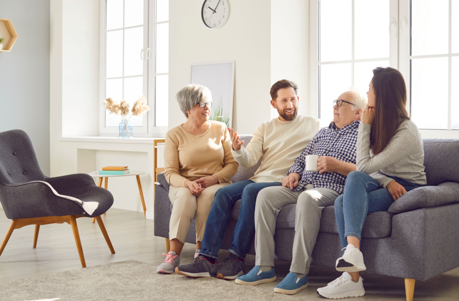 Four people sitting on a couch in a sunlit room having a cheerful group conversation over coffee.