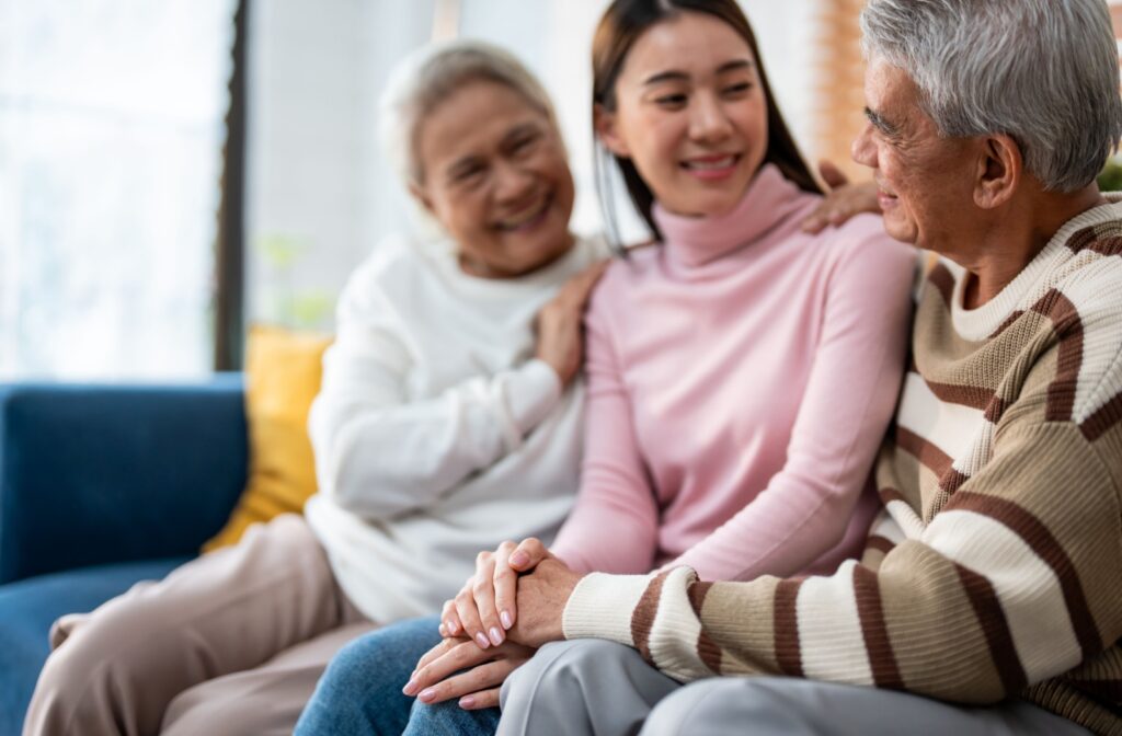 Adult with senior parents sitting closely on a couch smiling and talking while holding hands in a supportive moment.