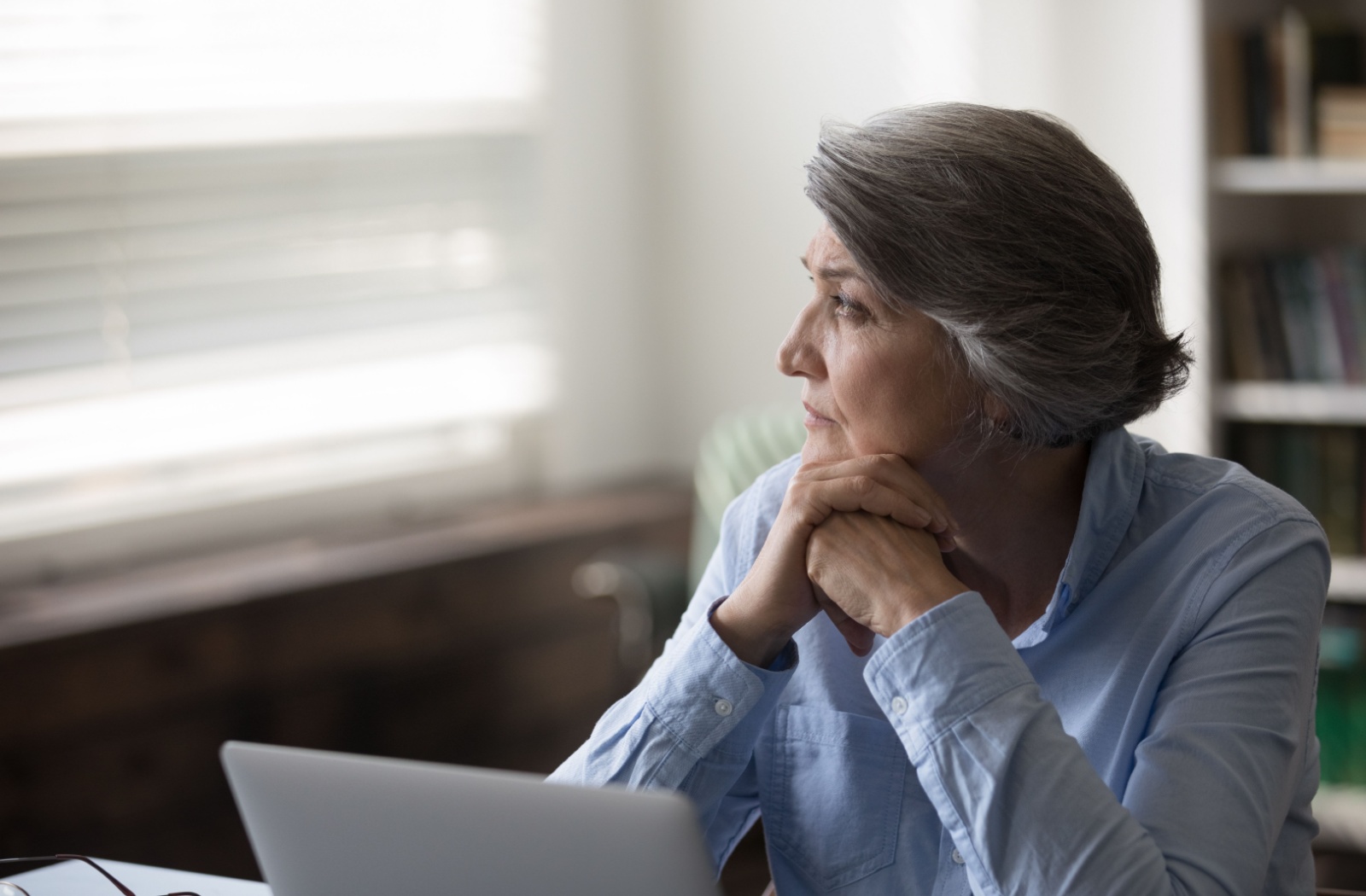 Older adult sitting at a desk with hands under chin gazing out a window with a thoughtful expression.