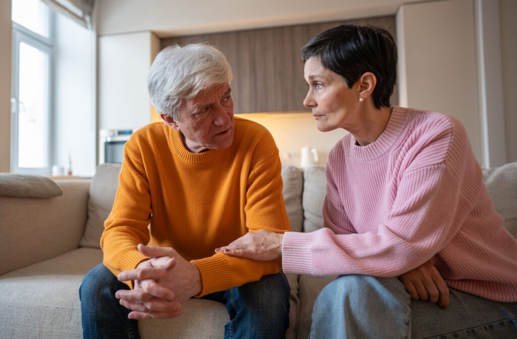 Two people sitting on a couch having a serious conversation while one offers comfort with a hand on the other's arm.