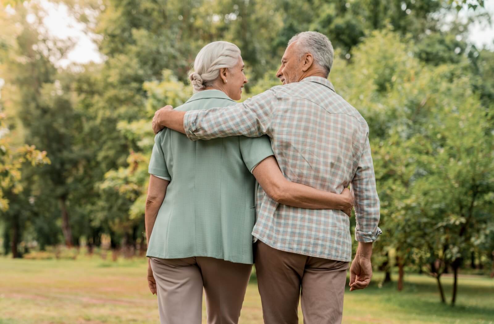 A shot from behind of a healthy older couple walking through a park and smiling at one another.