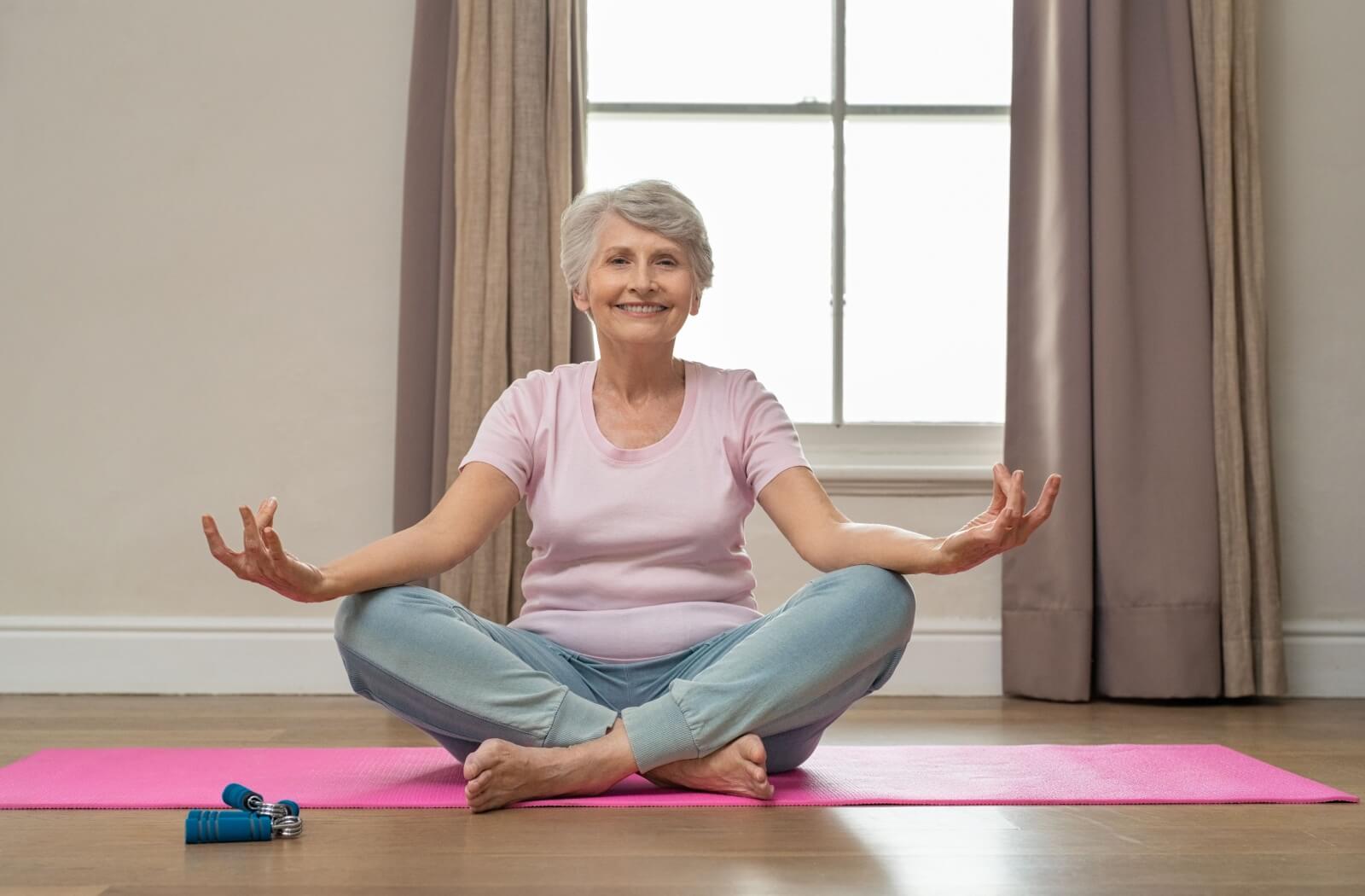 An older adult meditating at home on a pink yoga mat and smiling.