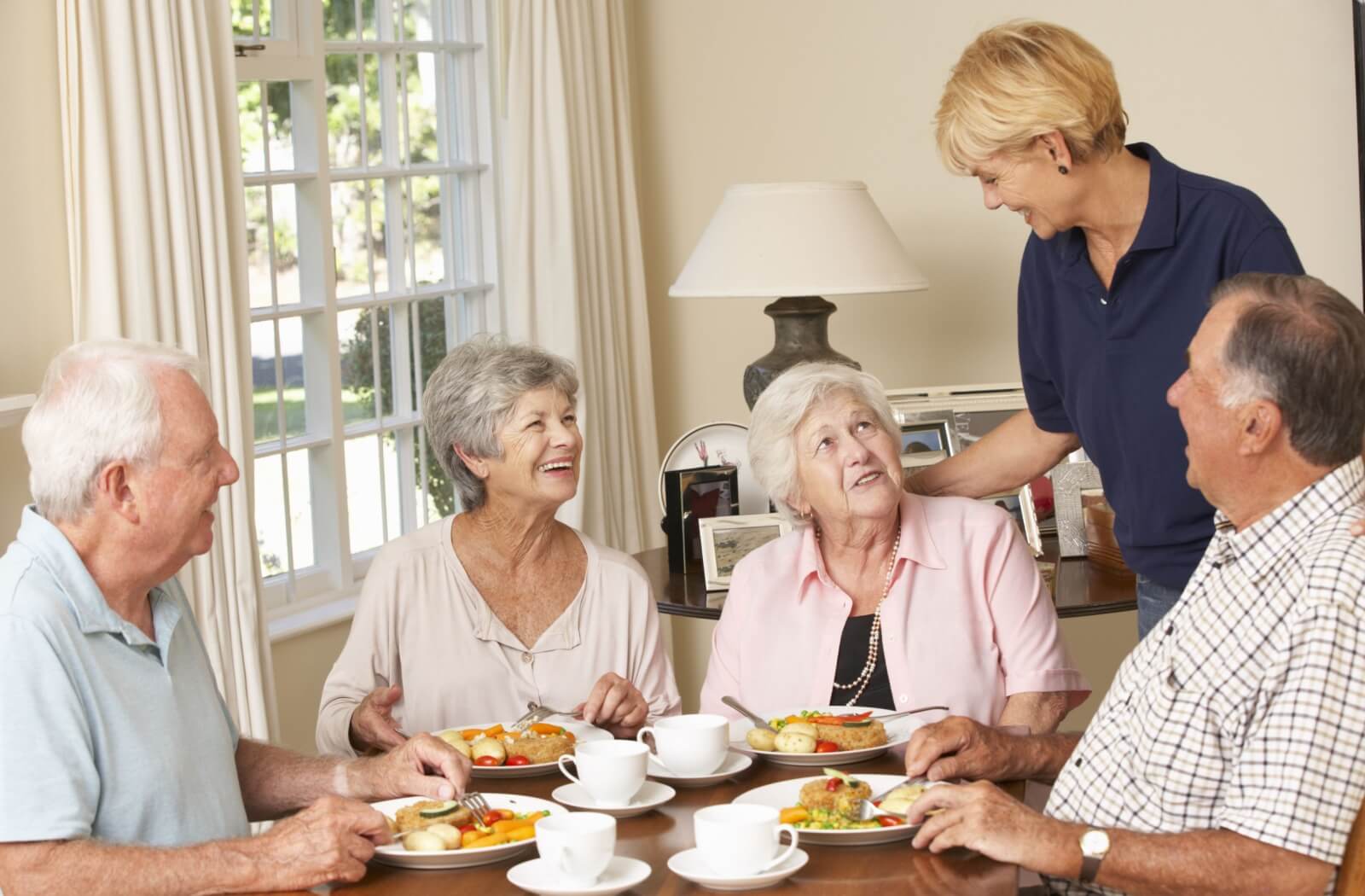 A grorup of residents look up at a caregiver and smile during a checkin while eating a group breakfast together in senior living